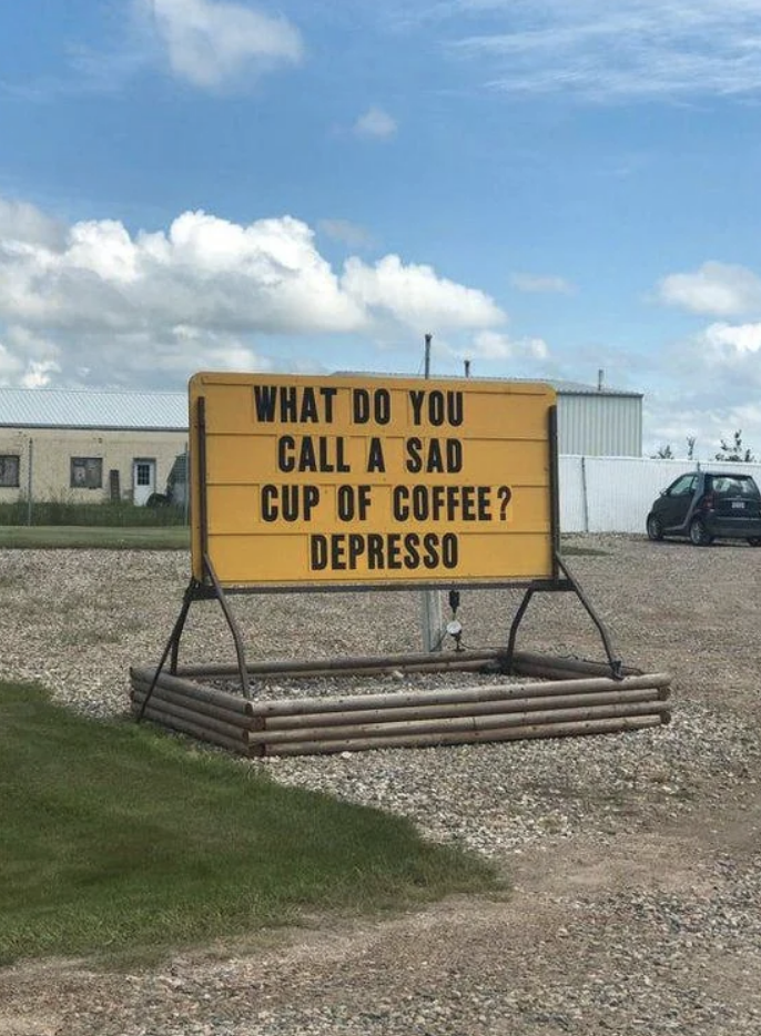 Sign with joke: &quot;WHAT DO YOU CALL A SAD CUP OF COFFEE? DEPRESSO&quot; displayed outside, with a grass field and building in the background