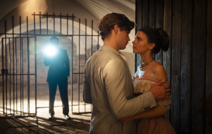 Dos personajes se miran íntimamente en un escenario dramático, con una persona al fondo iluminada por una luz blanca