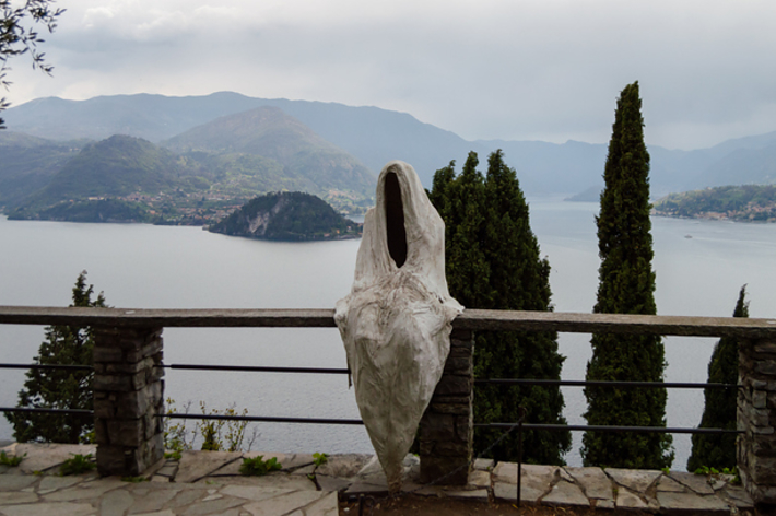 Sculpture of a figure with no head or arms overlooking a lake with mountains and cloudy sky