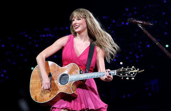 Taylor Swift performs onstage with acoustic guitar, wearing a pink dress