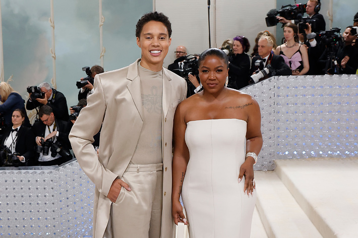 Two individuals posing at an event, one in a beige suit, the other in a white strapless dress. Photographers in the background