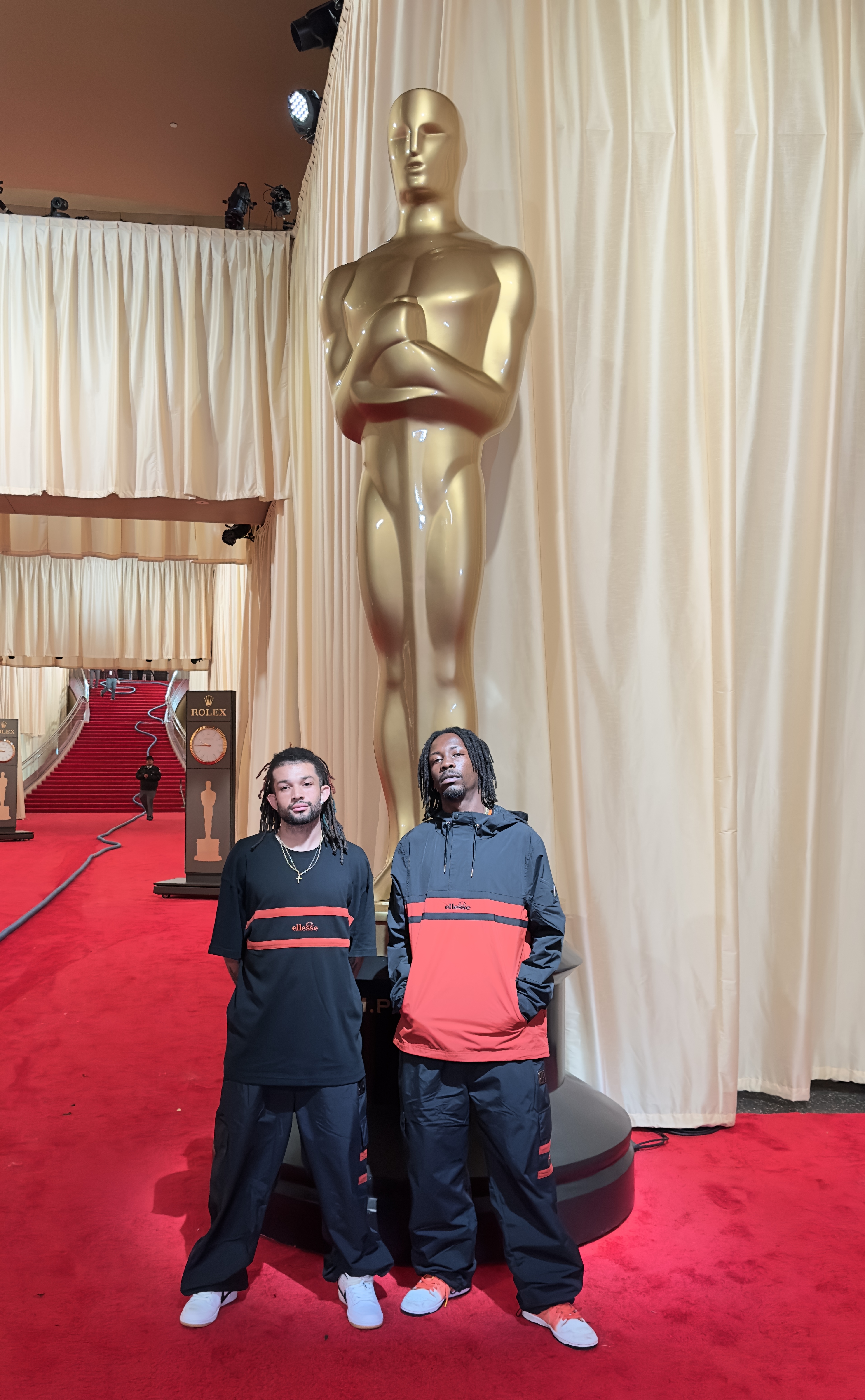 Two individuals in casual black and red attire standing in front of a large Oscar statue on a red carpet