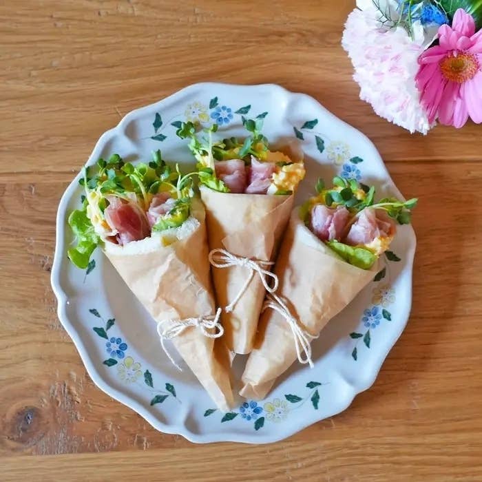 三つのハムとサラダのサンドイッチが花柄の皿に盛られていて、隣にはピンク色の花がある。
