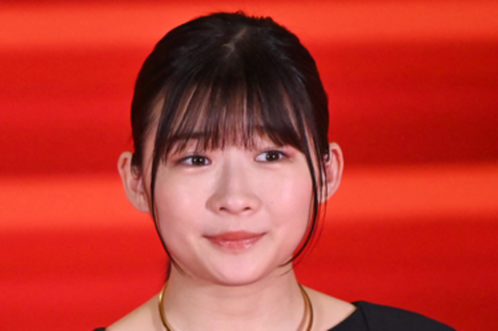 レッドカーペットに立つ女性、黒いドレスを着てカメラに向かって笑顔で手を振る。