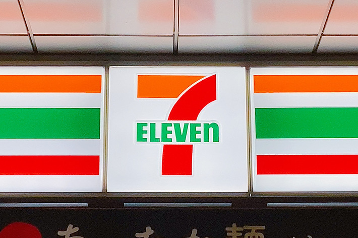 セブンイレブンの店舗看板、ロゴと照明付きの文字、下に日本語で商品宣伝のテキスト。