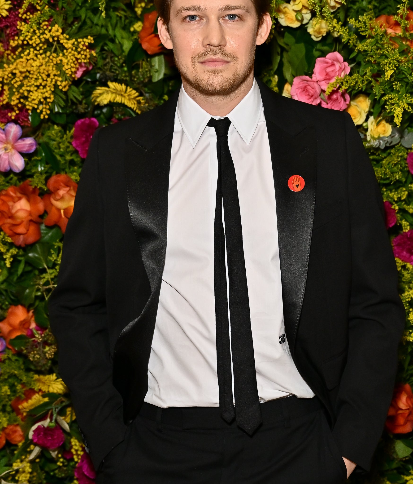 Joe Alwyn standing in front of a floral backdrop wearing a black suit with a white shirt and black tie