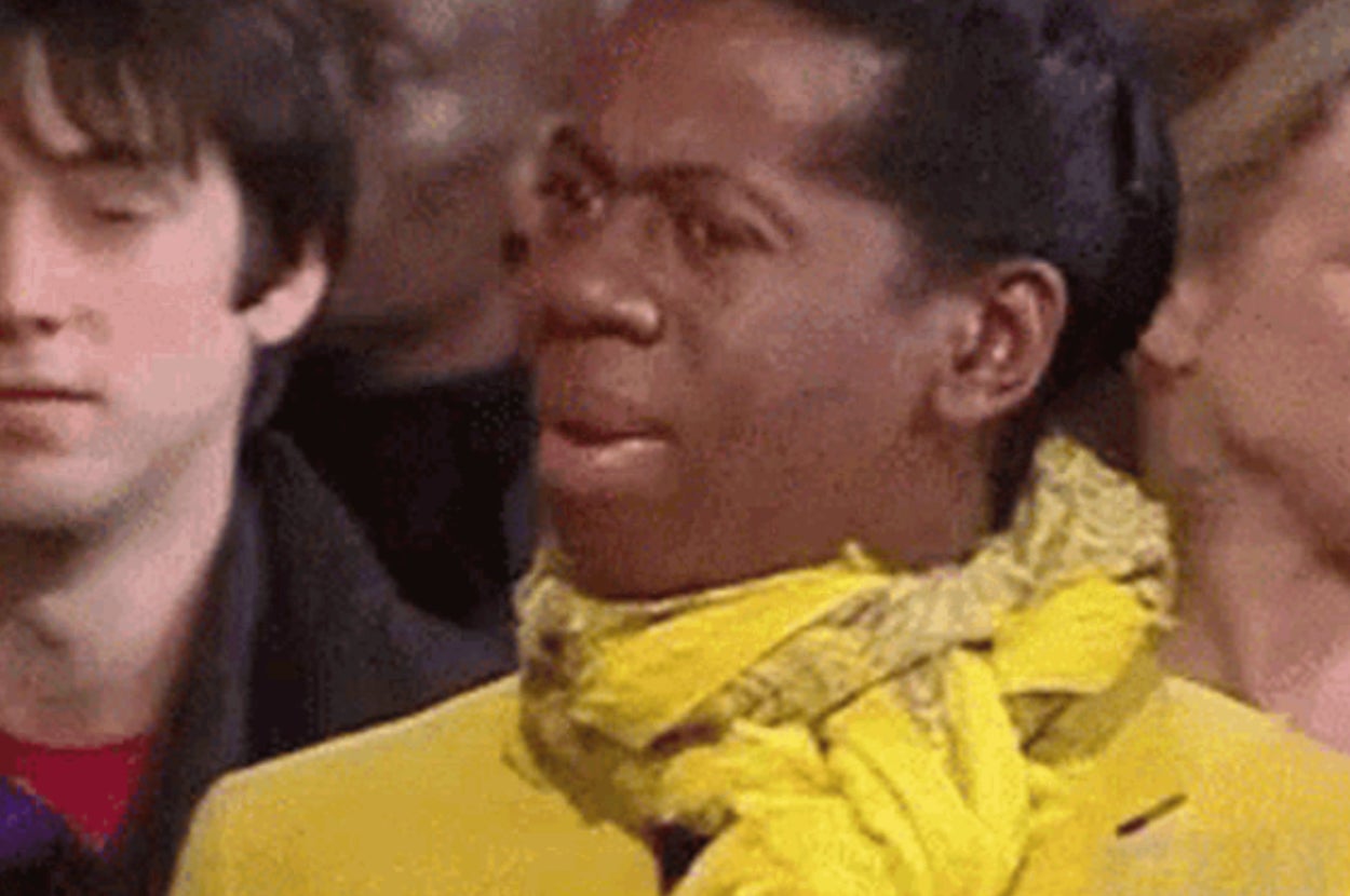 A person wearing a yellow scarf looks surprised among others in a crowd scene from a TV show