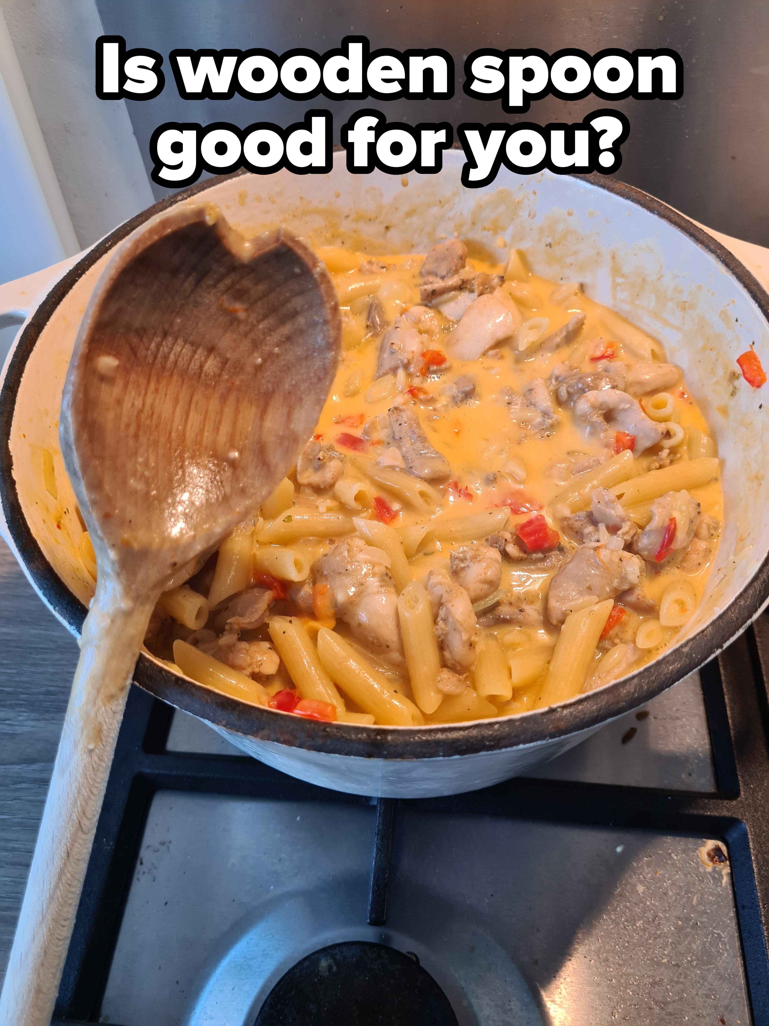 A pot of pasta with creamy sauce, chicken pieces, and red bell peppers on a stove. A wooden spoon rests inside the pot