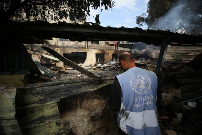 A person in a UNRWA vest stands amid the debris of a damaged building, inspecting the area