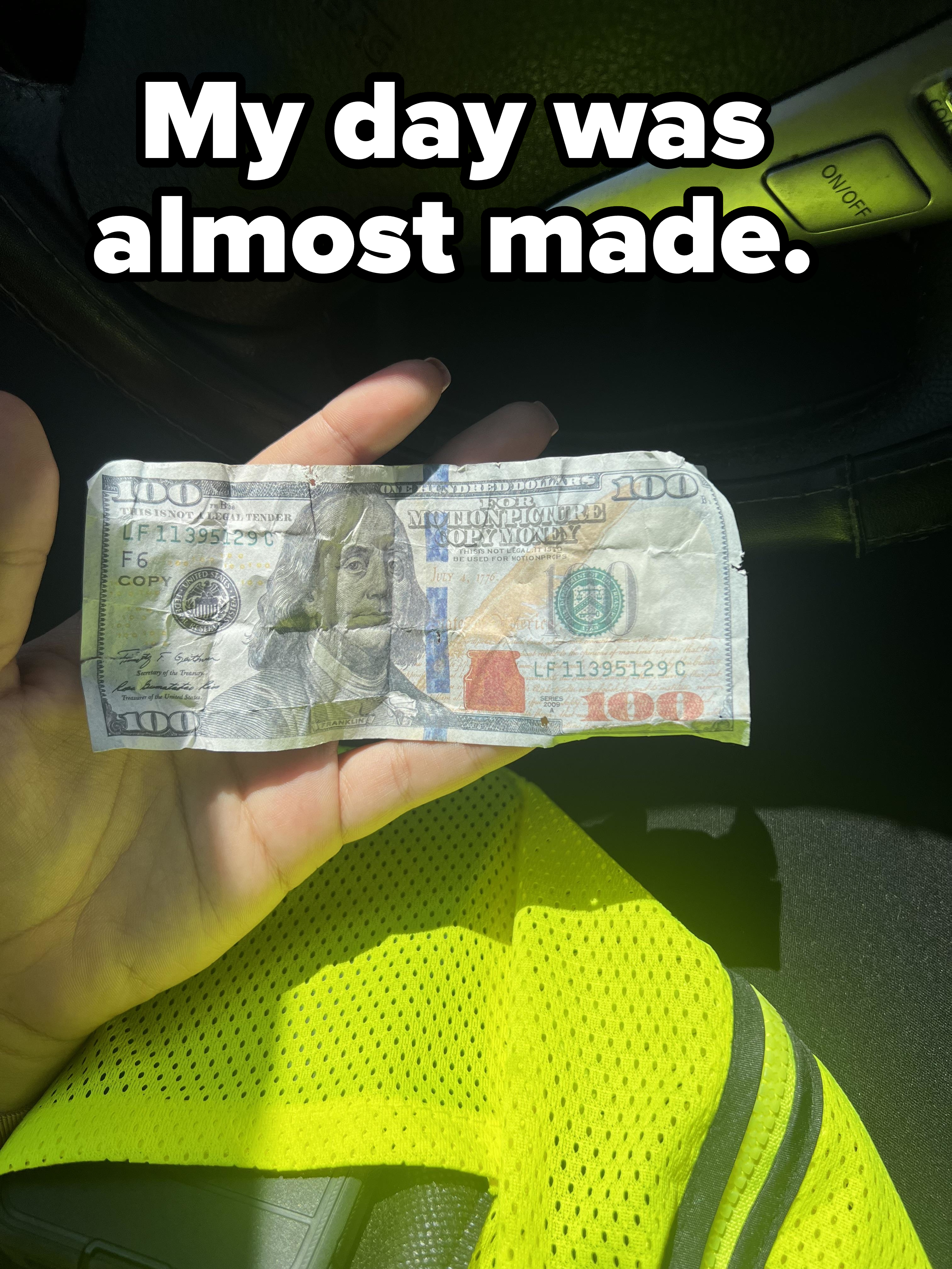 A hand holding a crumpled $100 bill inside a car with part of a yellow high-visibility vest visible underneath