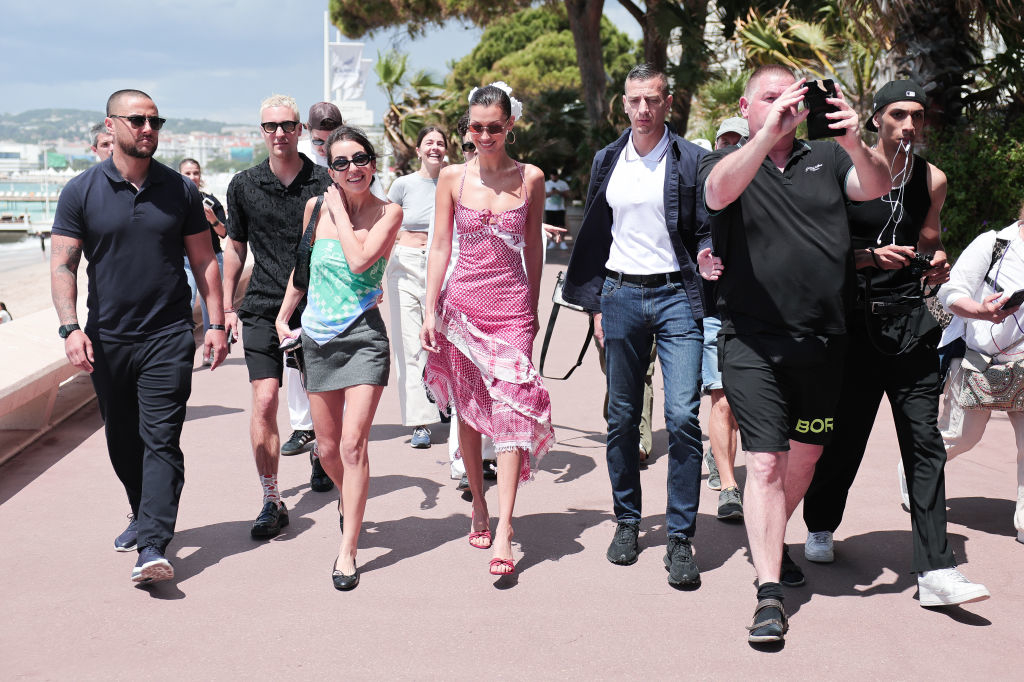 Selena Gomez walks along a promenade with a group of people, wearing a sleeveless dress with a pattern and sunglasses. An entourage surrounds her