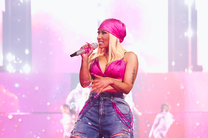 Cardi B performs on stage in a pink top, ripped jeans, and a pink headscarf, holding a microphone. Pink, ethereal background and backup dancers visible