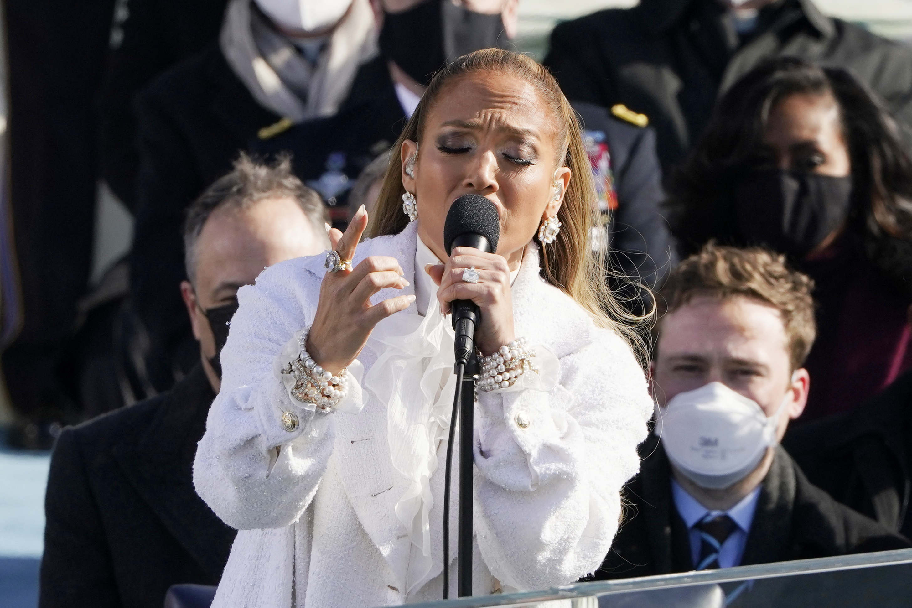 Jennifer Lopez sings into a microphone while wearing a white outfit and pearl accessories at an outdoor event. People are visible in the background