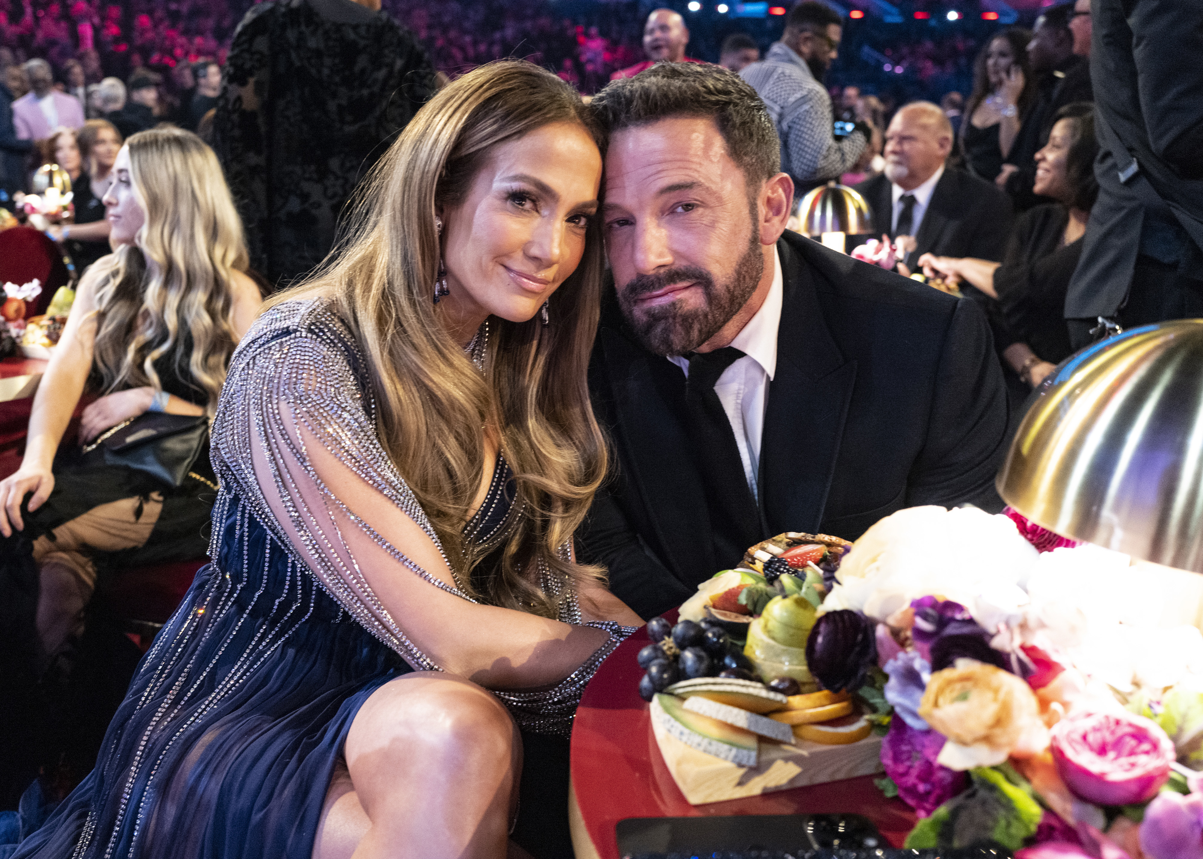 Jennifer Lopez in an elegant, sequined dress posing next to Ben Affleck in a suit at an event, seated at a table with a cheese and fruit platter