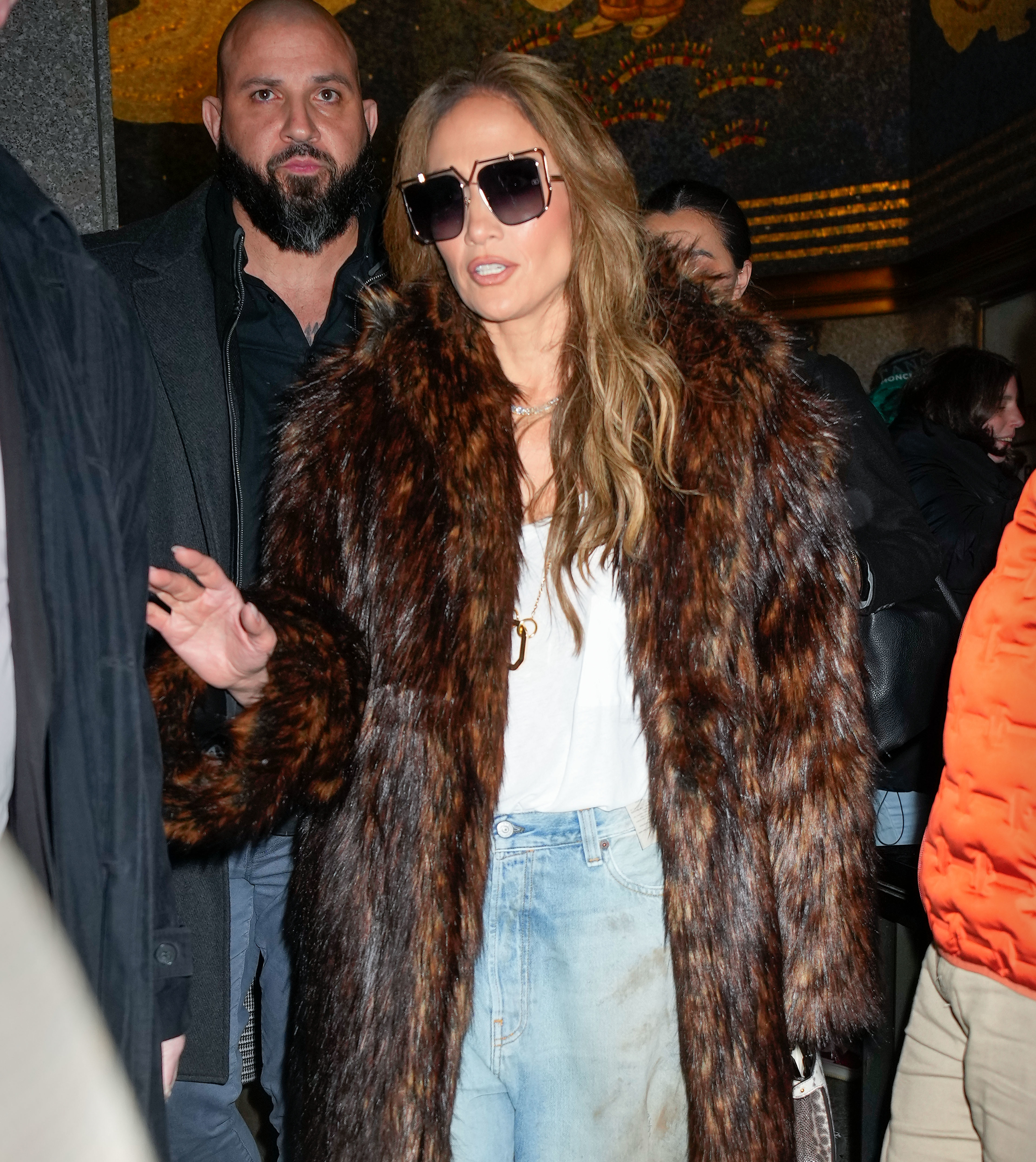 Jennifer Lopez wearing sunglasses, a white shirt, blue jeans, and a long fur coat, walking outside with a crowd and a security guard nearby