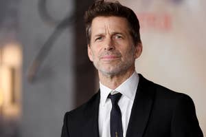 Zack Snyder on the red carpet, wearing a black suit jacket, white dress shirt, and black tie, looking into the distance