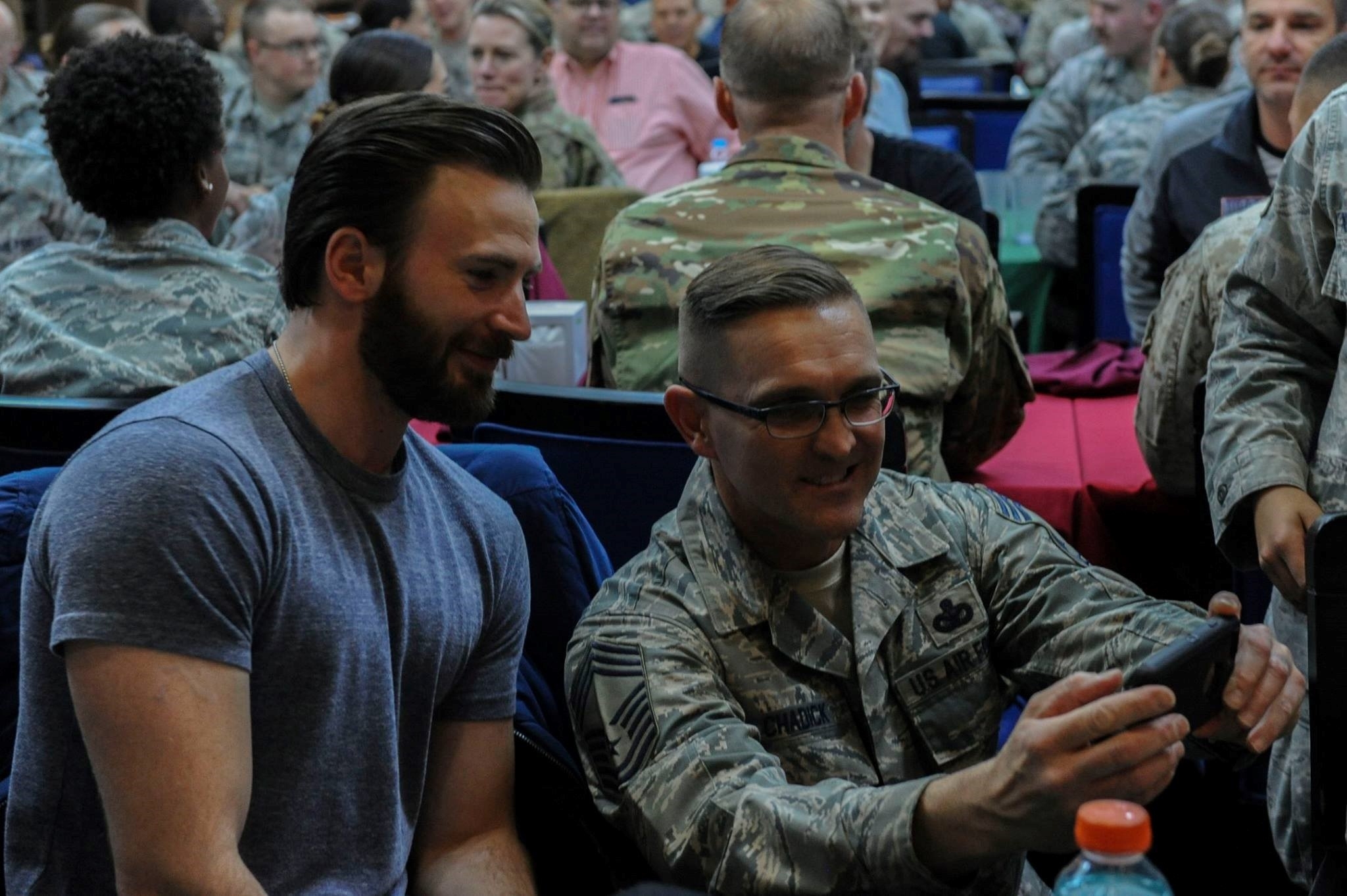 Chris Evans takes a selfie with a service member in uniform at an event surrounded by people, some in military attire and some in civilian clothes