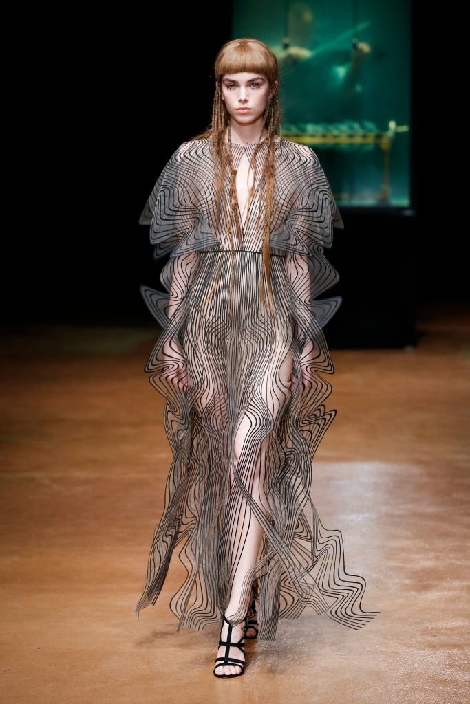 A model walks the runway wearing a sheer geometric dress with intricate patterns and braided hair at a fashion show