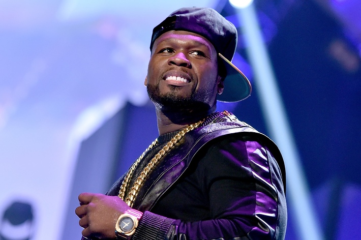 50 Cent on stage wearing a black leather-sleeved jacket, multiple gold chains, a wristwatch, and a backward baseball cap. He is smiling
