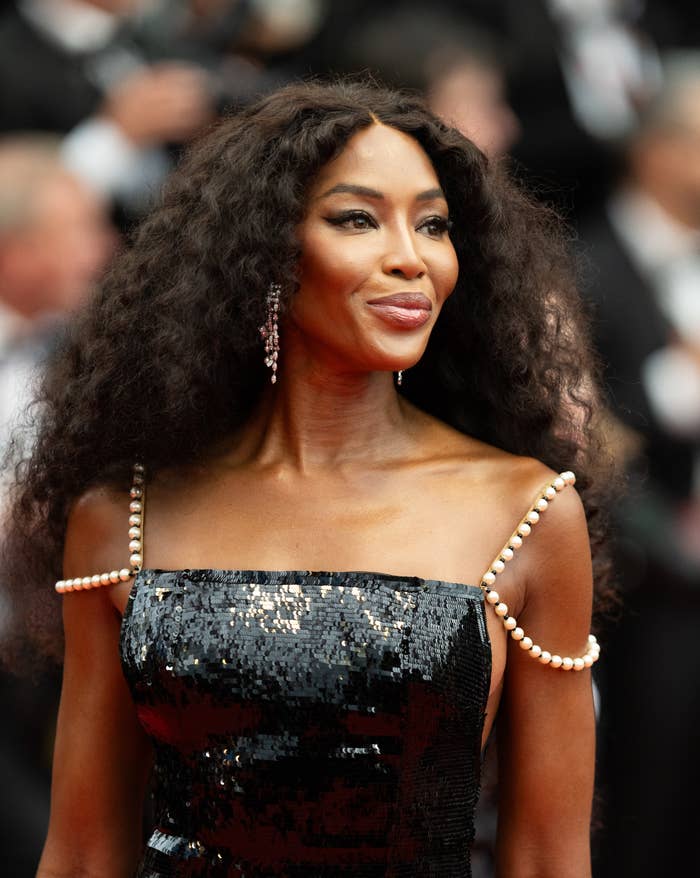 Naomi Campbell on a red carpet wearing a sequined dress with pearl shoulder straps, smiling at the camera