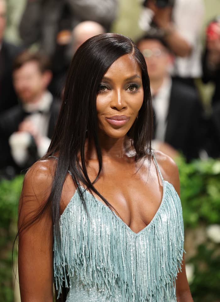 Naomi Campbell on the red carpet, wearing a spaghetti-strap dress with shimmering fringe details, photographers and attendees in the background