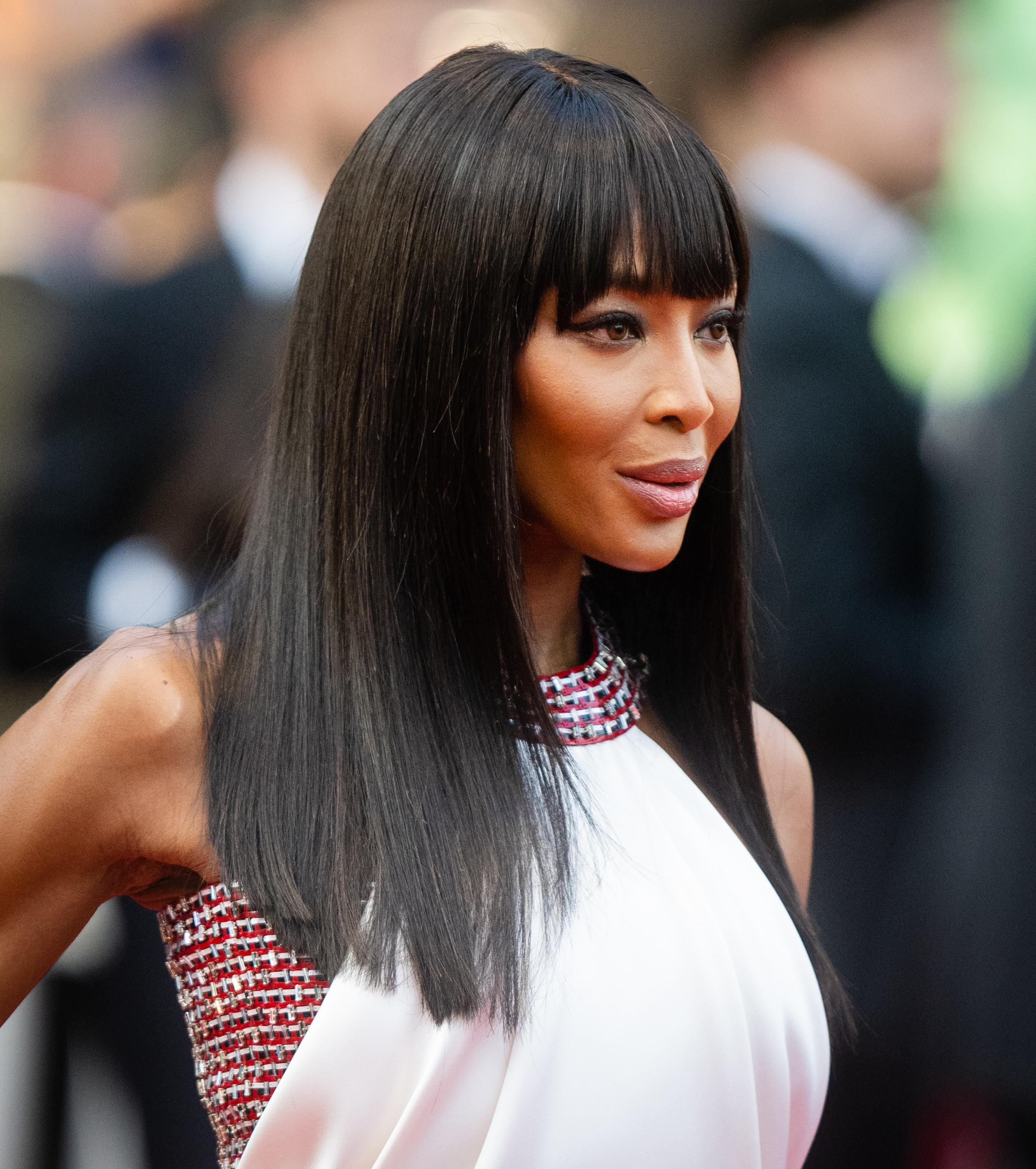 A woman poses on a red carpet, wearing a sleeveless gown with a high neckline and fringe detailing