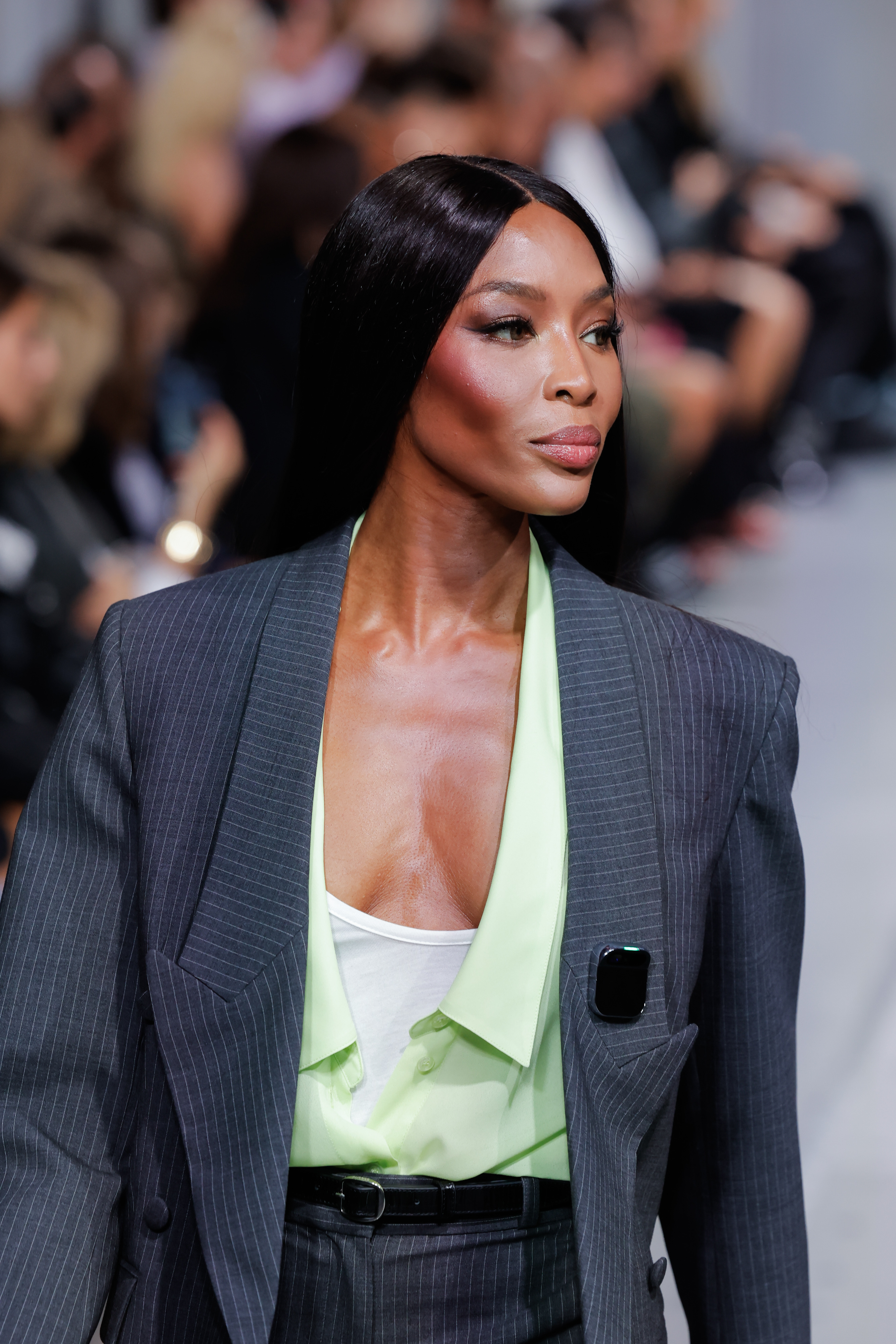 Naomi Campbell in a pinstripe blazer over a brightly-colored blouse with a plunging neckline as she walks a runway