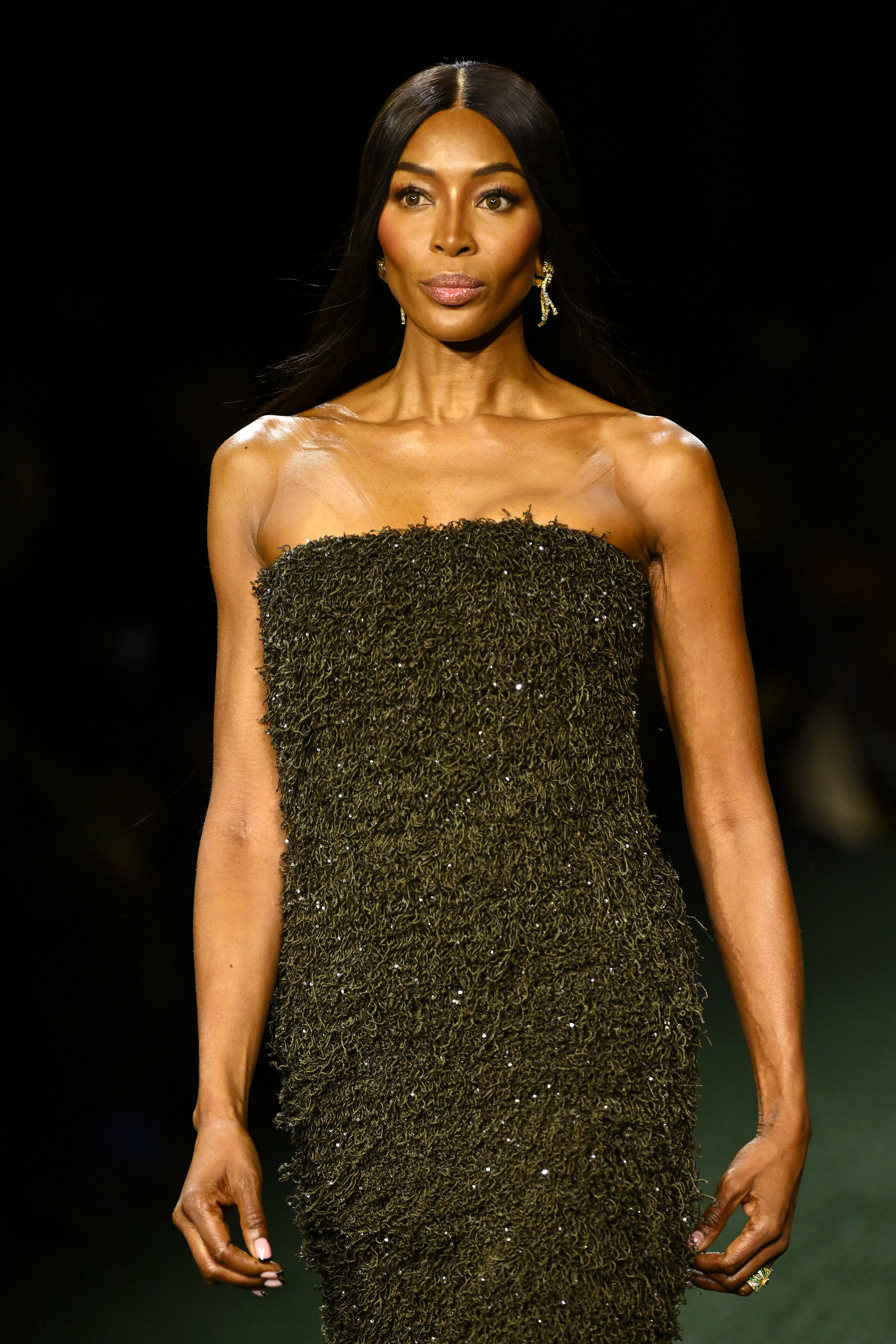 Naomi Campbell in a strapless, textured dress on a runway. Her hair is long and straight, and she wears dangling earrings