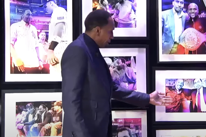 Stephen A. Smith pointing at a wall of framed photos featuring various moments with individuals, including one with a boxing champion belt