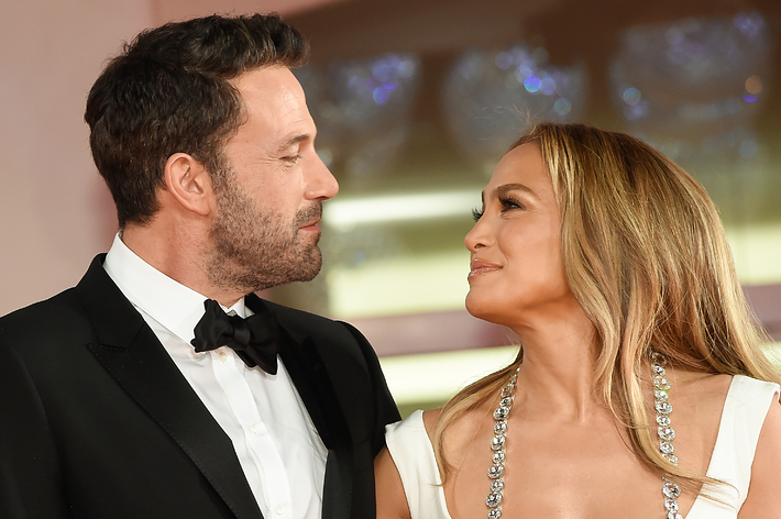 Ben Affleck in a tuxedo and Jennifer Lopez in a low-cut white dress with a sparkling necklace, smiling at each other on a formal red carpet event