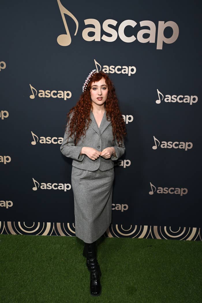 Chappell Roan with long curly hair wearing a suit, skirt, and boots poses in front of an ASCAP backdrop
