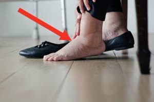 A person is holding their swollen ankle and foot, with one black flat shoe on the floor beside them. The person is seated and appears to be in discomfort