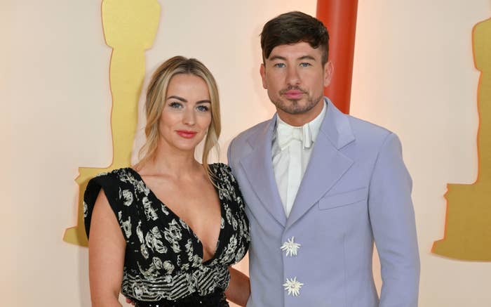 Barry Keoghan and Alyson Sandro at an event posing and wearing stylish outfits. Barry wears a light-colored suit with a shirt and bow tie, and Alyson is in a low-cut dress