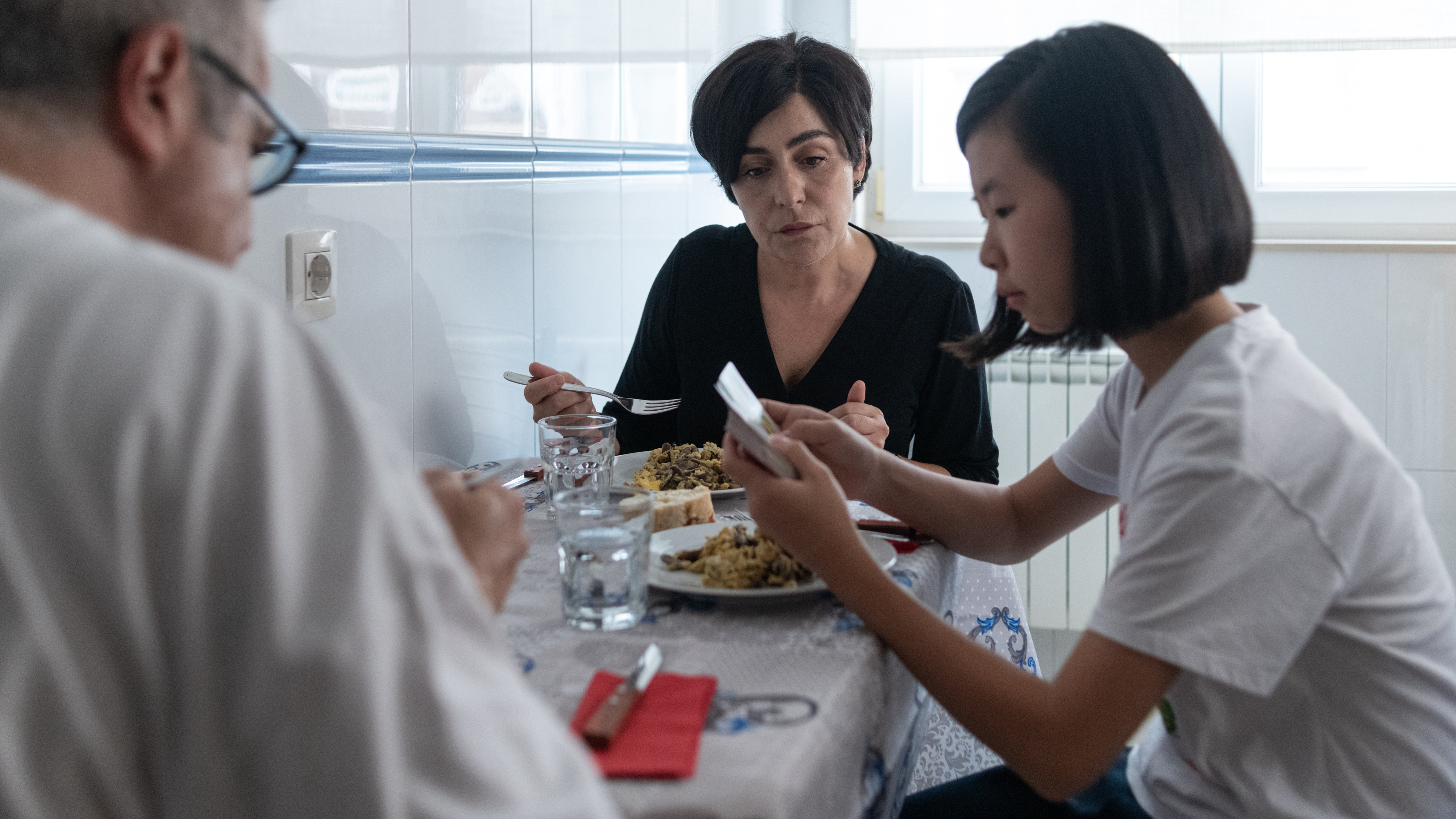 People eating at a kitchen table, with a focus on the young woman with shoulder-length hair on the right looking at her phone. Names are unspecified