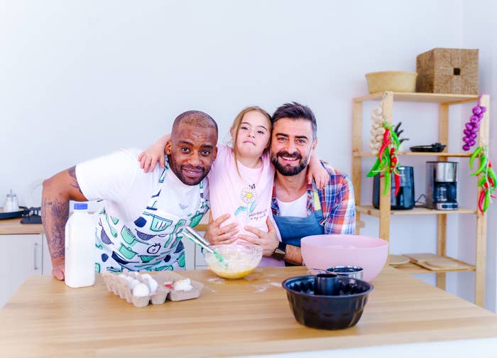Two men and a young girl with Down syndrome are smiling in a kitchen, preparing to bake with ingredients and utensils on the table