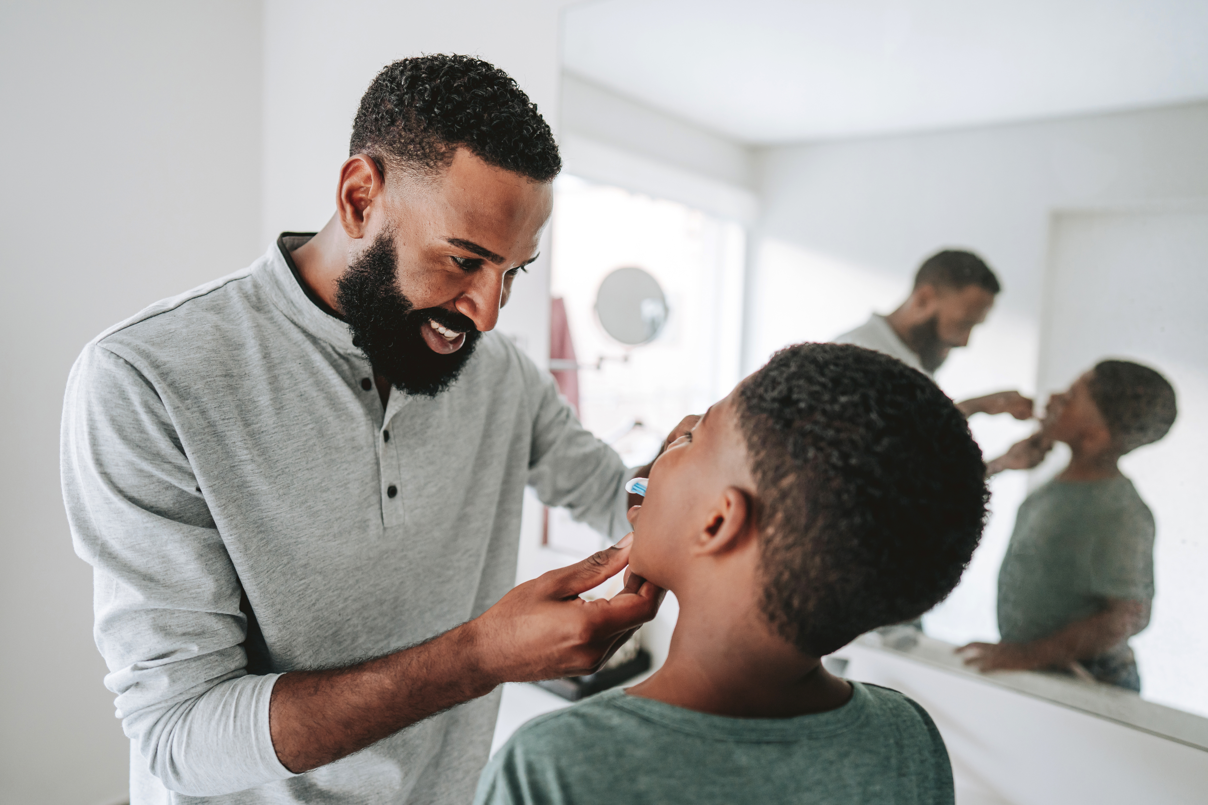 A man shaves a younger boy's face with a razor in front of a bathroom mirror. Both are smiling and enjoying the moment