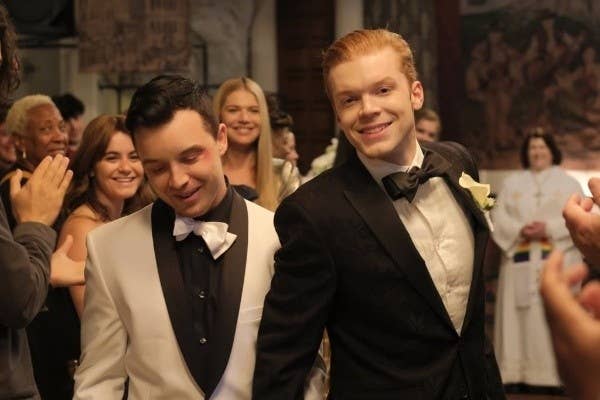 Ian y Mickey de Shameless. Viste dos hombres en traje, uno en blanco y otro en negro, sonríen en lo que parece ser una celebración, con gente aplaudiendo detrás