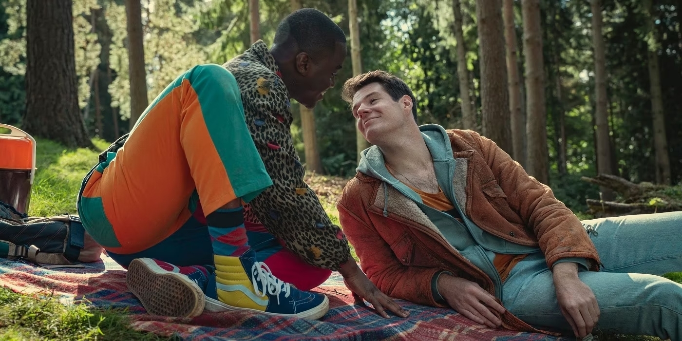Dos personas sentadas en un picnic en el bosque, una vistiendo ropa colorida con chaqueta de leopardo, y la otra con chaqueta marrón y suéter gris