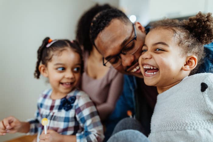 A man smiling and happily playing with two young girls, one in a checkered dress holding a lollipop, and the other in a light top