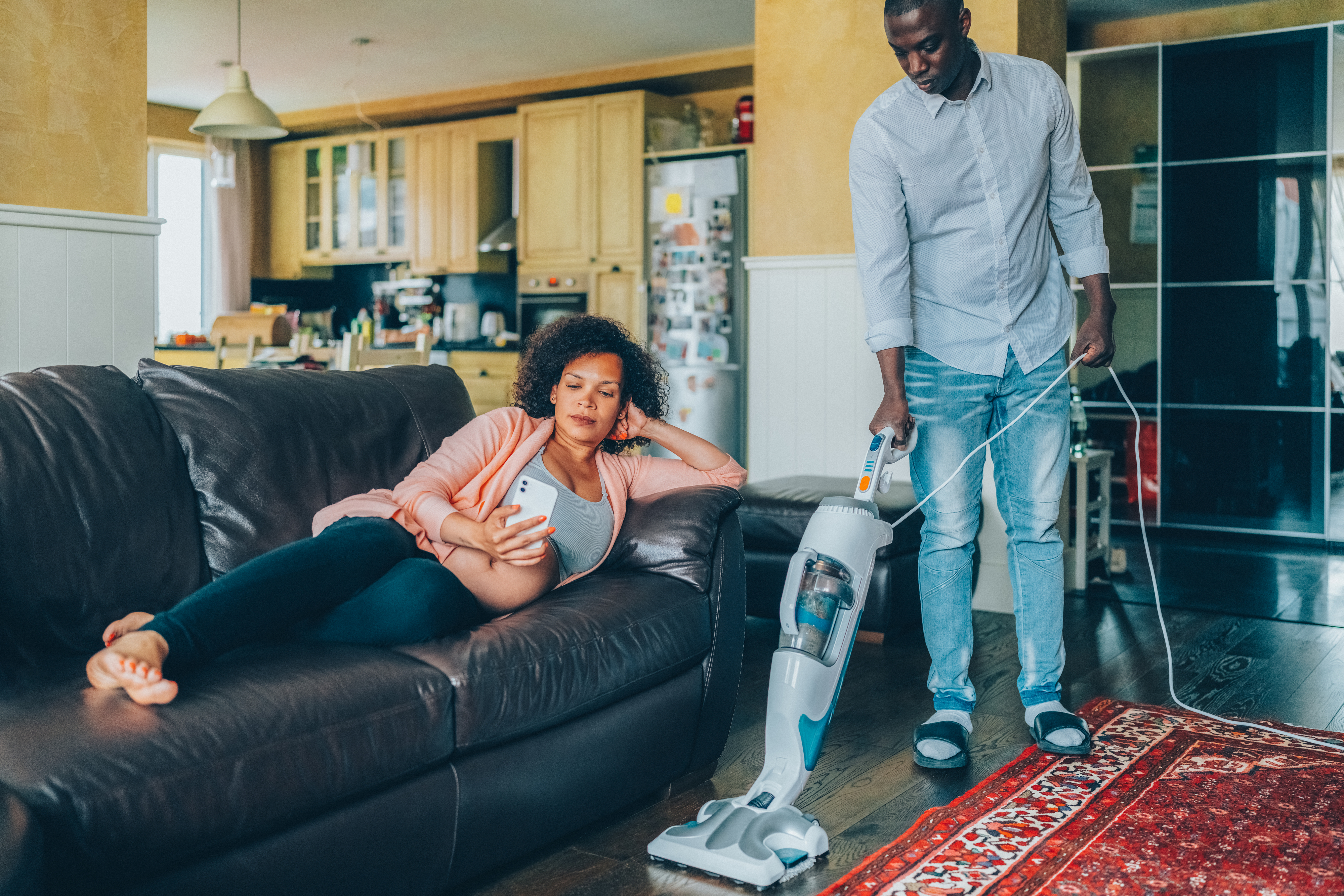 A pregnant woman reclines on a couch looking at her phone while a man vacuums the living room nearby