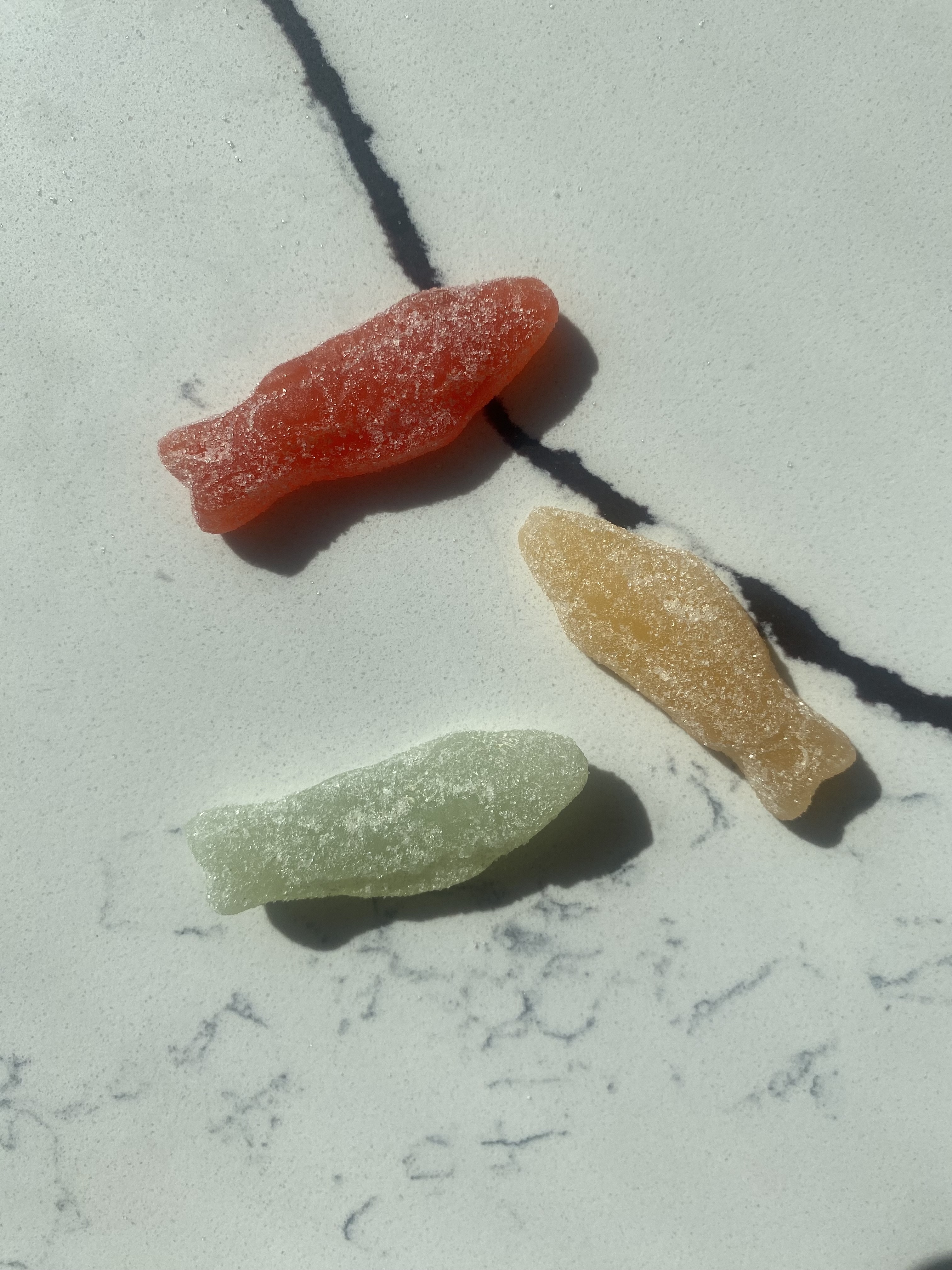 Three sour gummy fish candies displayed on a marble surface, one red, one green, and one yellow