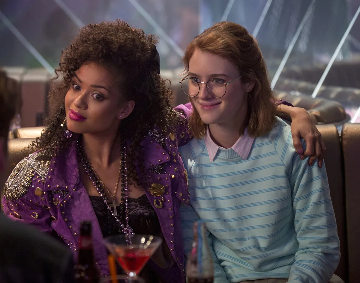 Gugu Mbatha-Raw con chaqueta de estilo punk y Mackenzie Davis con suéter a rayas, abrazadas en un bar con bebidas sobre la mesa