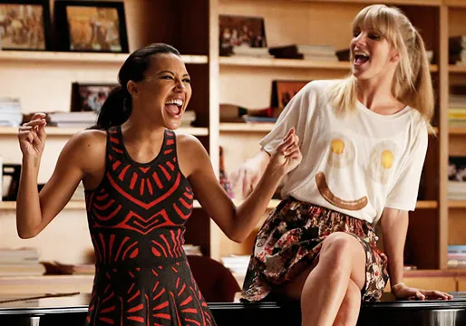 Naya Rivera y Heather Morris reíen juntas. Naya lleva un vestido estampado y Heather una camiseta con diseño y una falda estampada. Están en un ambiente casual