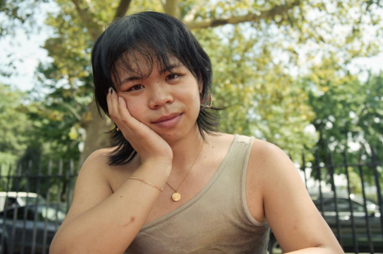Person wearing a tank top and necklace, resting their head on their hand, outdoors in a park-like setting. Trees and a fence are visible in the background