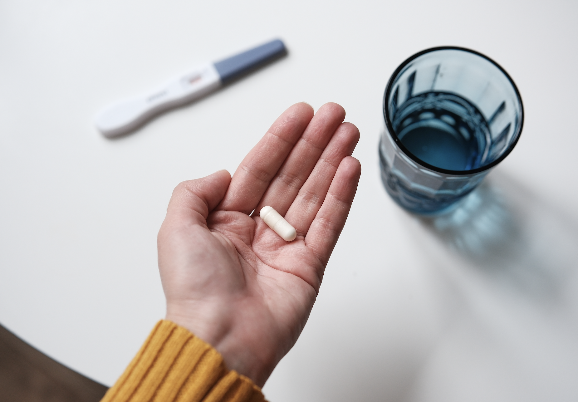 A hand holding a pill and a capsule with a pregnancy test and a water glass in the background