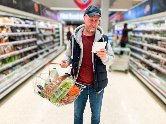 A man in casual attire, including a cap and hoodie, is holding a shopping basket full of groceries in a supermarket aisle, reading a list