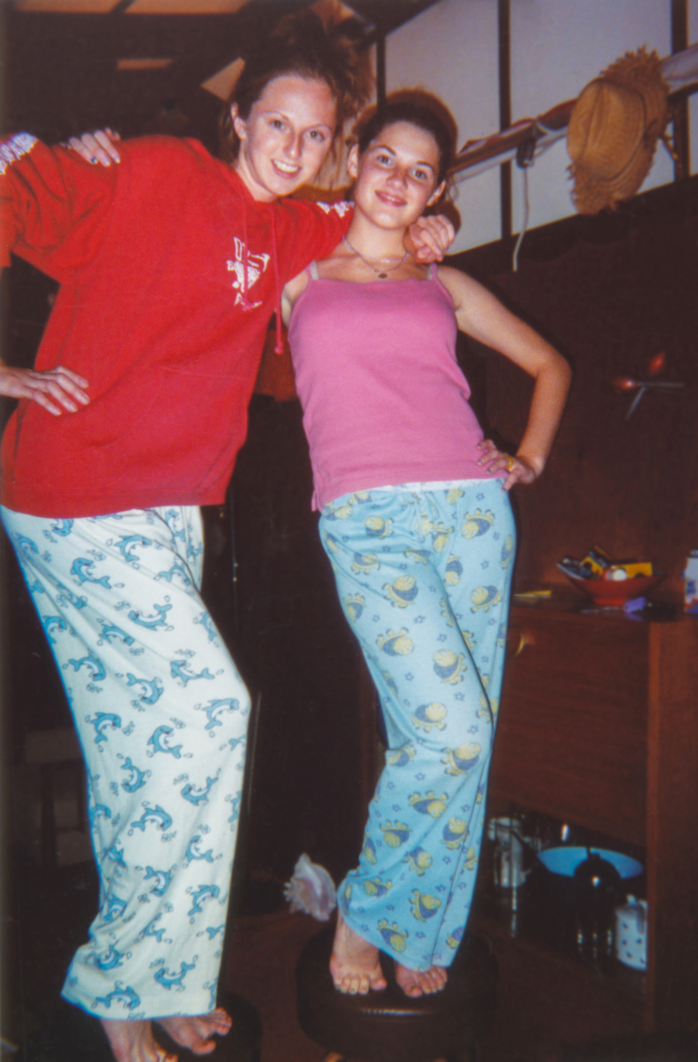 Two people, name unknown, posing and smiling in pajamas inside a room. One wears a red sweatshirt and patterned pants, the other a pink tank top and pajama pants