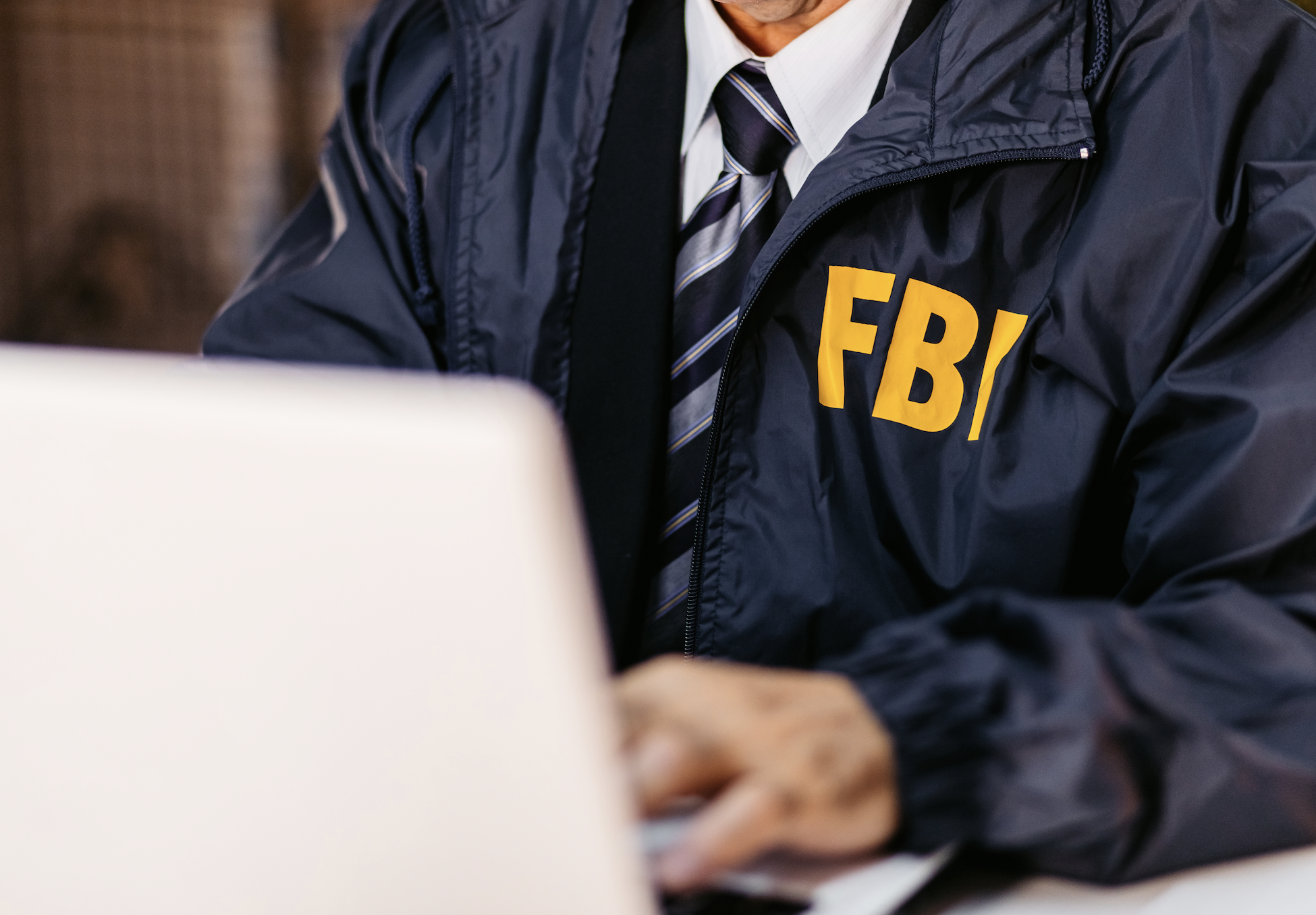 An FBI agent in a jacket with "FBI" in yellow letters works on a laptop