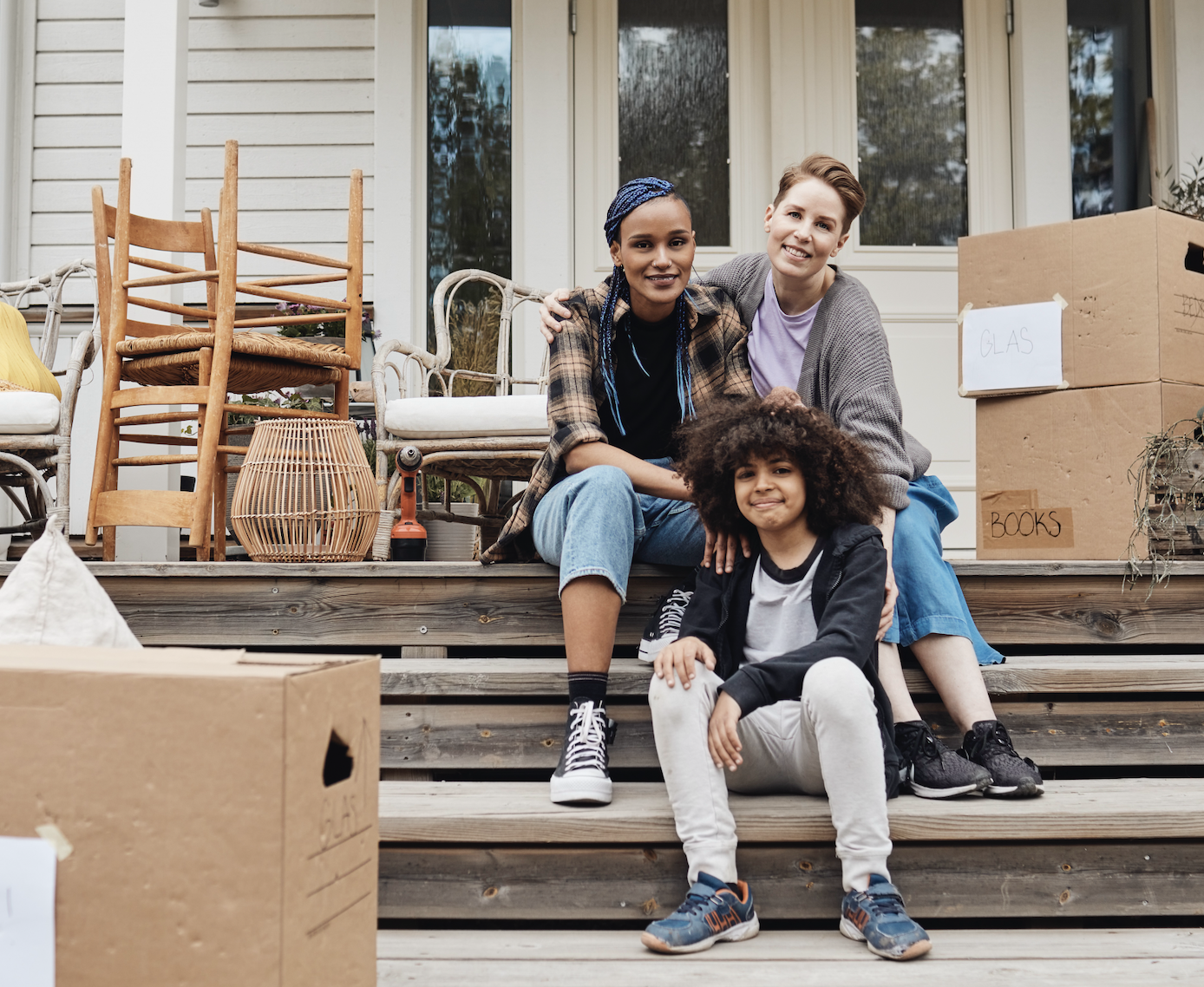 Two adults and their child sit on the steps of a house surrounded by moving boxes and chairs. They are smiling and appear relaxed