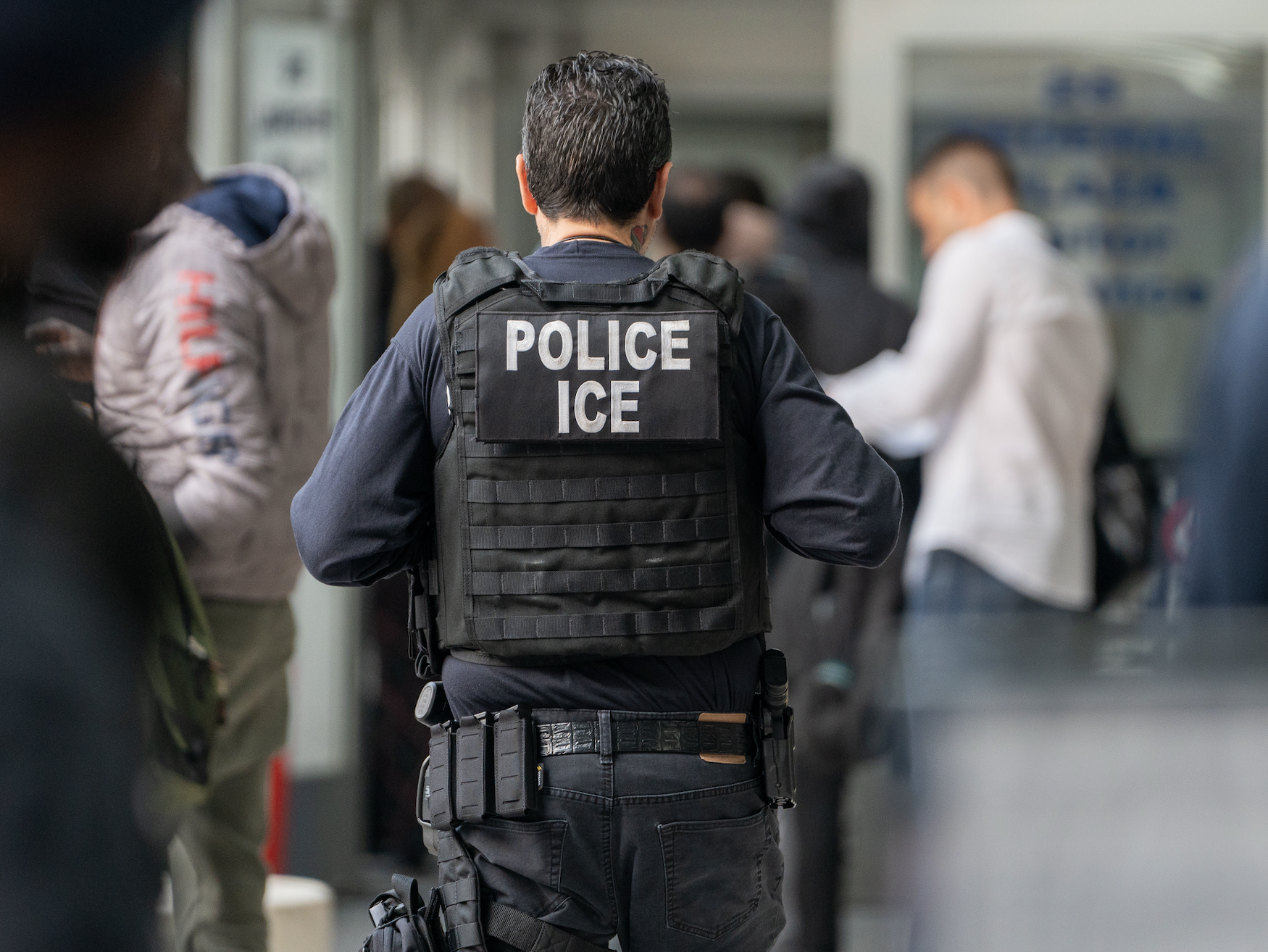 A police officer with "POLICE ICE" on his back stands in a busy public area. Many people are around, some blurred and walking past